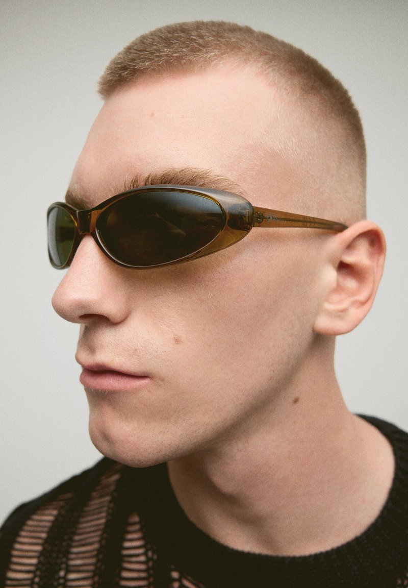 Young person with short hair wearing narrow, dark oval sunglasses and a black textured top, looking slightly to the left.