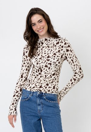 Donna sorridente che indossa una maglia beige a maniche lunghe con macchie marroni animalier e jeans blu con tasche borchiate su sfondo semplice.