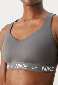 Siva sportska grudnjak s V-izrezom, izrađen od glatkog materijala, s bijelim Nike logotipom na elastičnoj traci.