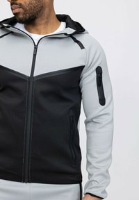 Veste de sport avec fermeture éclair, présentant un schéma de couleurs noir et gris, finition mate, capuche et poches zippées sur les manches.