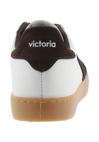 Zapatilla blanca con un acento de ante marrón en el talón. Presenta una suela de goma con un detalle en color arena cosido. El nombre de la marca "victoria" aparece en el talón.