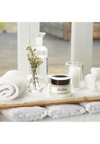 AHAVA Active Moisture Gel Cream i en klar glasburk med brunt lock, bredvid en glasvas med gröna och vita blommor samt vita handdukar.
