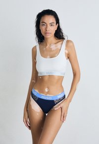 White cropped tank top with "HUGO" branding in gray underband, paired with navy high-waisted briefs featuring a blue polka dot band.