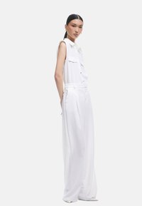 Haut sans manches blanc avec des détails de boutons et deux poches avant, associé à un pantalon blanc taille haute à jambes larges en tissu lisse.
