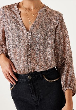 Nainen, jolla on kuvioitu pitkähihainen paita, jossa on napit, mustat korkeavyötäröiset farkut, joissa metalliset koristukset taskuissa, sekä kerrokselliset kultaiset kaulakorut.