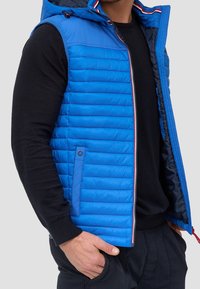 Chaleco acolchado azul con capucha, diseño de rombos horizontales, cierre de cremallera y acentos en rojo y blanco. Se lleva sobre un suéter de punto negro.