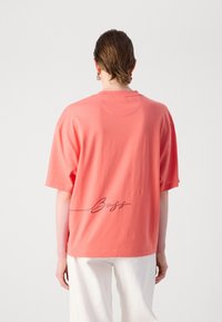 Coral oversized cotton t-shirt with short sleeves and a round neck. Features a cursive “Boss” logo printed on the back. Plain texture.