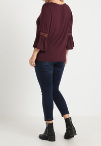 Viininpunainen paita levenevillä hihoilla ja pitsikoristelulla, yhdistetty tummiin farkkuihin ja mustiin nilkkureihin. Sileä kangaspinta.