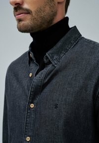 Camisa de mezclilla gris oscuro con cuello abotonado, que presenta botones de madera y un pequeño logotipo bordado en el lado izquierdo. Tejido texturizado.