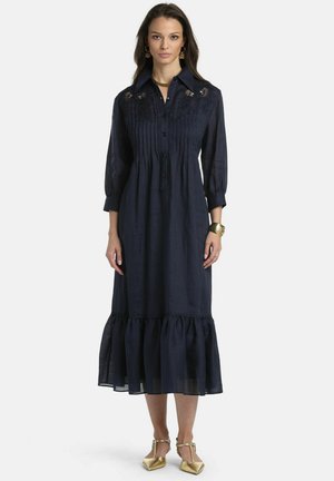 Kvinde stående iført en navyblå midi-kjole med 3/4-ærmer, blonder og plisserede detaljer, flæsekant, guldbøjle, øreringe og guldsko.