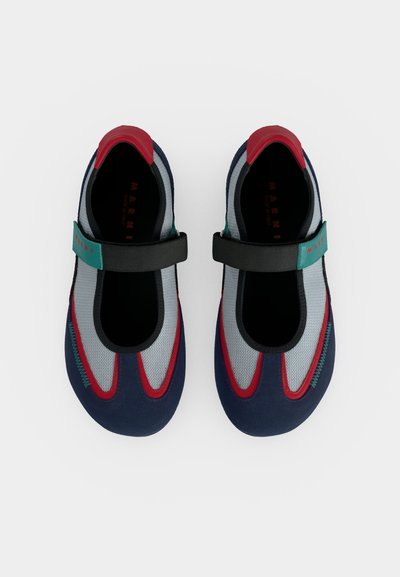 Paire de chaussures style Mary Jane à bout fermé en tissu bleu marine, rouge et gris avec une bride noire avec un patch vert sur fond blanc.