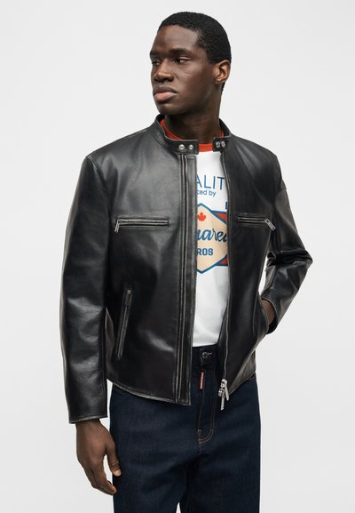 Άνδρας με μαύρο δερμάτινο μπουφάν πάνω από λευκό μπλουζάκι με γραφικά και σκούρο τζιν, στέκεται με το ένα χέρι στην τσέπη, κοιτώντας αριστερά.