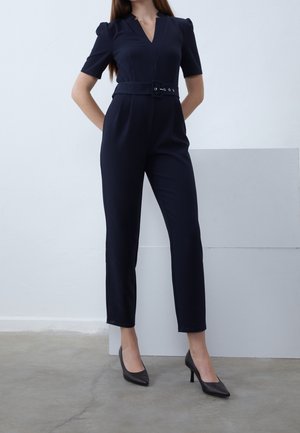 Vrouw draagt een zwarte jumpsuit met riem en korte mouwen en zwarte hoge hakken, staand tegen een effen lichte achtergrond.