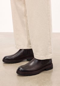 Bocanci din piele maro, cu o finisare netedă, cu panouri laterale elastice și o talpă texturată, asociați cu pantaloni bej pe un podea deschisă.