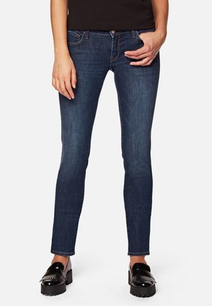 Femme portant un jean slim bleu foncé, des mocassins noirs à plateforme, debout devant un fond uni, les mains près des poches.