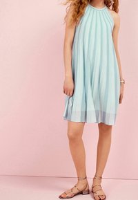 Vaaleansininen ilman hihoja oleva mekko, jossa on pystysuorat raidat, korkea kaula-aukko ja laskeutuva siluetti. Yhdistetty metallisiin hihna-sandaaleihin.