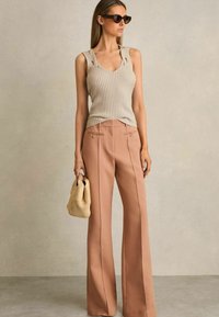 Top de punto acanalado beige sin mangas con cortes en los hombros, combinado con pantalones de pierna ancha rosa claro, que presentan pliegues frontales y lazos laterales, sosteniendo un bolso de mimbre.
