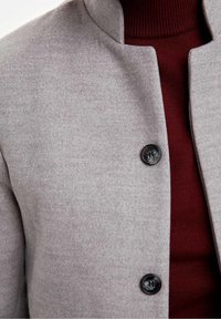 Graue Jacke aus Wollmischung mit hohem Kragen, glatt strukturiert, mit einem schwarzen Knopfdetail. Kombiniert mit einem gerippten burgunderroten Rollkragenpullover darunter.