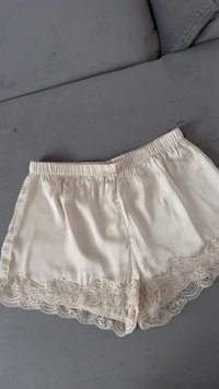 Cream satin shorts with an elastic waistband and decorative lace trim at the hem, featuring a smooth texture and relaxed fit.