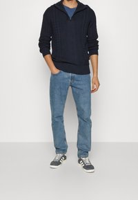 Marinblå stickad tröja med halvdragkedja, prydd med flätstickade mönster, i kombination med ljusblå jeans och mörka sneakers med vita snörningar.