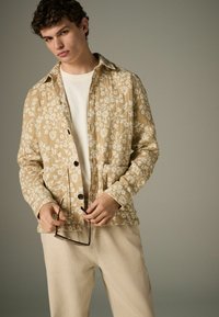 Jachetă cu un model floral bej deschis, cu guler, închidere cu nasturi și două buzunare frontale, purtată peste o cămașă albă și pantaloni deschiși.