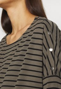 Top riscado verde azeitona com linhas horizontais pretas. Apresenta um decote redondo e detalhes em botões metálicos nos ombros. Textura de tecido suave.