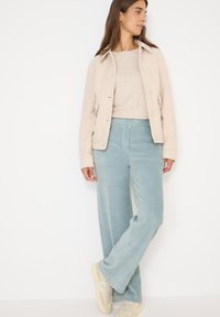 Beżowa kurtka z sztruksu z detalami guzików, połączona z jasnobeżowym swetrem i jasnooliwkowymi spodniami z szerokimi nogawkami także wykonanymi z sztruksu. Całość dopełniają casualowe sneakersy.