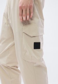 Μπεζ παντελόνι cargo από ελαφρύ ύφασμα, με πλαϊνή τσέπη και μαύρο τετράγωνο λογότυπο με τη λέξη «BOSS» με πράσινα γράμματα.