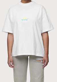 Bílé oversized tričko z bavlny s kulatým výstřihem. Obsahuje zelené a žluté vyšívané logo na hrudi. Nosené s světle šedými potrhanými džíny.