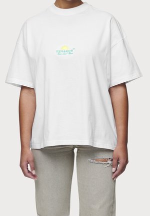 Camiseta blanca de algodón de gran tamaño con cuello redondo. Presenta un logo bordado en verde y amarillo en el pecho. Se lleva con unos jeans rasgados en gris claro.
