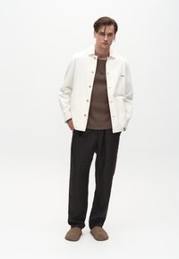 Hvid knap-op jakke over en brun tekstureret sweater, parret med løse sorte bukser og brune slip-on sko, stående foran en ensfarvet baggrund.