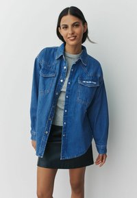 Denim shirt in blue, featuring two chest pockets, snap buttons, and a curved hem. Worn over a gray crewneck with a black leather skirt.