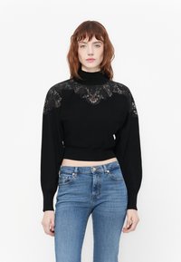 Pull noir court avec des manches longues et un col haut, présentant une dentelle sur les épaules et une texture côtelée au niveau de l'ourlet.
