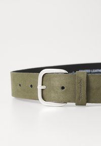 Ceinture en suède vert olive avec une boucle en métal rectangulaire argentée et un dos en cuir noir, présentant des trous uniformément espacés et une étiquette de logo.