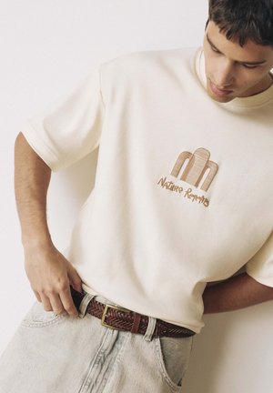 Junger Mann, der einen geflochtenen braunen Ledergürtel justiert, trägt ein hellbeiges T-Shirt mit "Nature Republic" Logo und hellfarbige Jeans.