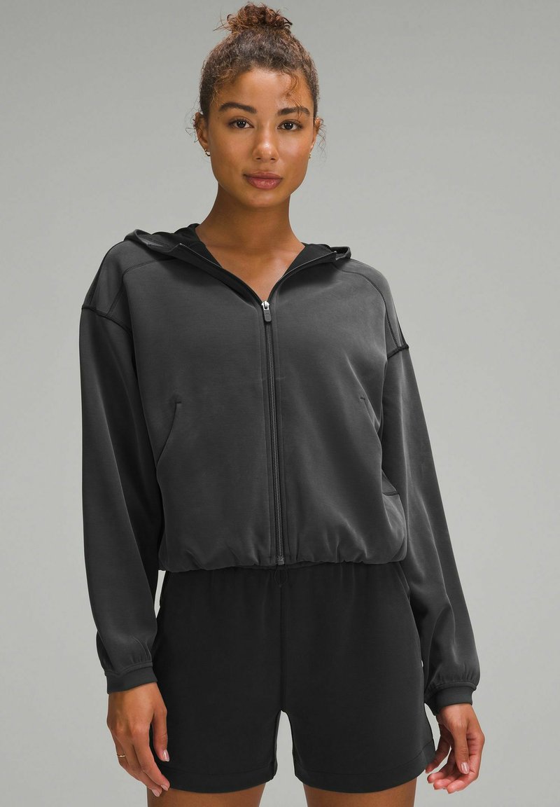 Black zip-up hoodie with a hood, featuring long sleeves, side pockets, and a cropped design, paired with black shorts. Smooth texture.