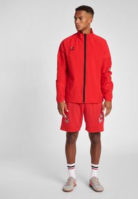 Rode sportjack met een hoge kraag en rits, gecombineerd met rode shorts met grijze zigzagpatronen. Draagt gestreepte sokken en lichte schoenen.