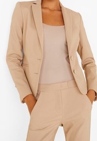 Beżowa marynarka o dopasowanym kroju z pojedynczym zapięciem na guzik, klapami i dwoma przednimi kieszeniami, noszona na dopasowanym topie bez rękawów i spodniami.