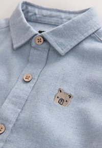 Camicia azzurro chiaro con colletto, in tessuto strutturato, con bottoni in legno e un ricamo a forma di orso sul lato sinistro.