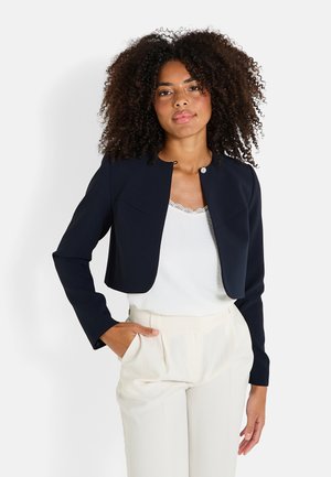 Veste cropped marine avec un col rond, des manches longues et un petit fermoir. Associée à un haut blanc et à un pantalon de couleur claire.
