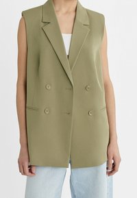 Blazer sans manches vert olive avec un design croisé, deux boutons et poches latérales. Assorti à un jean bleu clair.