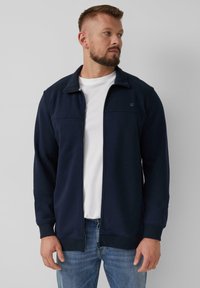 Marineblauwe zip-up jack met een hoge kraag, ribgebreide manchetten en contrasterende stiksels, gedragen over een wit t-shirt, in combinatie met lichtblauwe jeans.