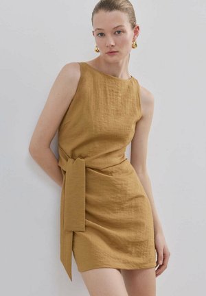 Femme portant une robe en lin moutarde sans manches avec ceinture à la taille, debout contre un mur clair uni, avec des boucles d'oreilles créoles dorées.