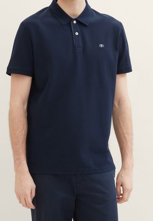 Homme portant un polo bleu marine à manches courtes avec deux boutons et un petit logo sur la poitrine gauche, associé à un pantalon bleu marine assorti.