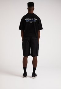 Schwarzes Baumwoll-T-Shirt mit grafischem Text auf dem Rücken, kombiniert mit knielangen schwarzen Cargo-Shorts mit Seitentaschen und strukturiertem Finish.
