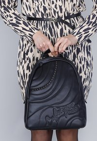 Zaino in pelle nera con design a spirale in rilievo, borchie argentate e logo. Dotato di manico superiore e chiusura con zip.