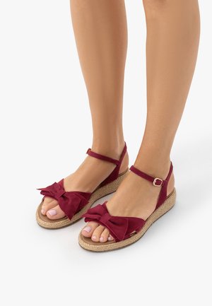 Nogi w czerwonych sandałach na kostkę z dużymi kokardami na plecionych koturnach, na jednolitym białym tle.