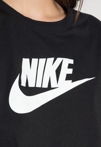 Černé tričko vyrobené z bavlny, s velkým bílým logem "NIKE" a designem swoosh na přední straně. Klasický kulatý výstřih.