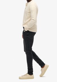 Top henley de manga larga de color beige claro con detalles de botones, combinado con jeans oscuros y zapatillas claras, mostrado de lado.