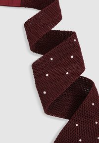 Fundă tricotată în nuanță de burgund cu o țesătură texturată, deschisă. Prezintă puncte albe distribuite uniform pe toată lungimea ei. Design flexibil și cu model.
